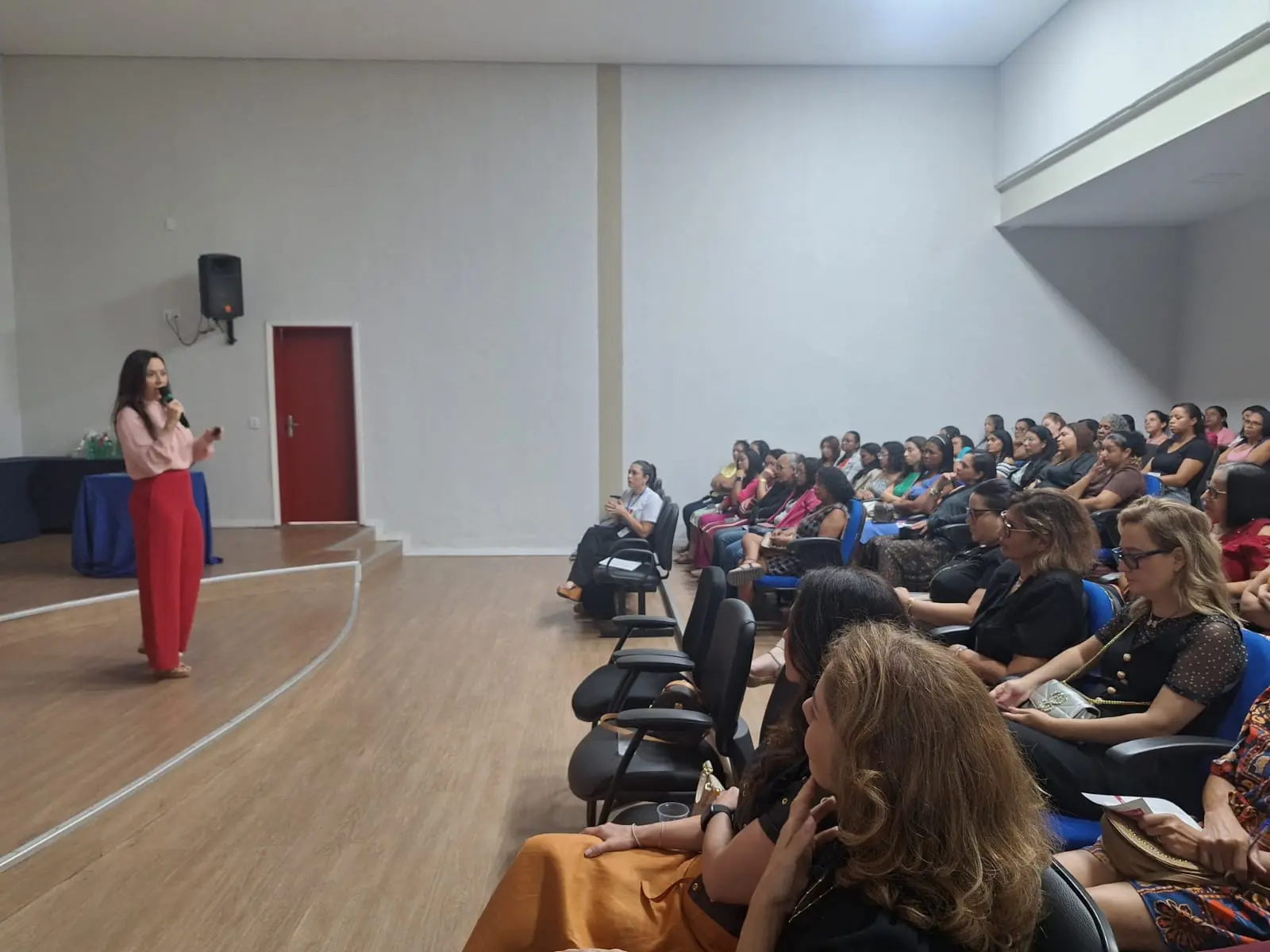 Foto horizontal em plano aberto que mostra a juíza Tatyana Lopes falando ao microfone, em pé, no auditório do fórum, para dezenas de mulheres que estão sentadas na plateia.